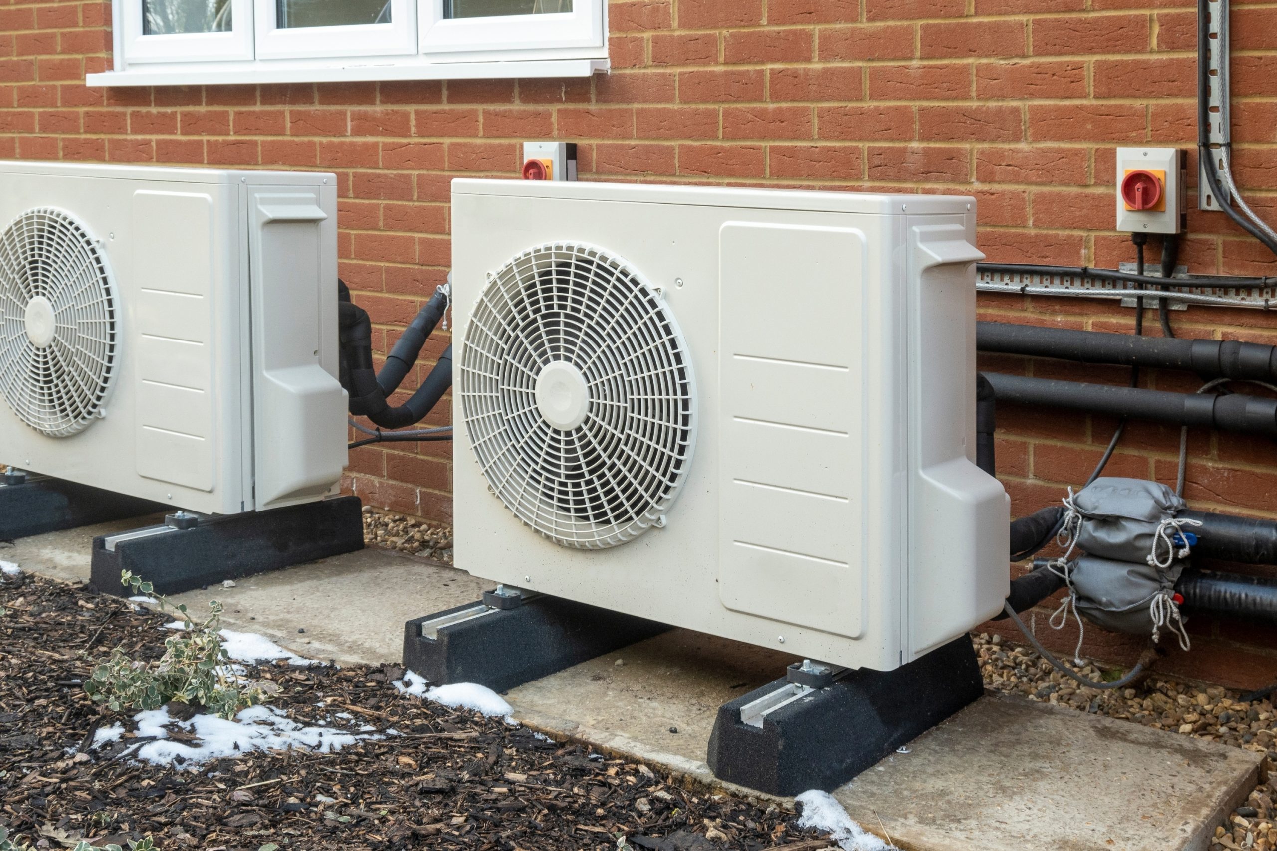 Two outdoor heat pump condenser units mounted on pads beside a brick wall